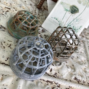 May include: Three vintage metal flower frogs in varying shades of gray, green, and brown. The frogs have a cage-like design with a circular base. A piece of rusty metal and a botanical print are in the background.