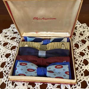 May include: An open vintage box with the text "Elgin American" contains five bow ties. The bow ties are various colors and patterns, including blue, gold, and red. The box is lined with a cream-colored fabric and has a red exterior.