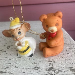 Può includere: Due piccole statuine in ceramica: un topo vestito da ape e un orsacchiotto. Il topo indossa un cappello giallo e un completo bianco con strisce nere e gialle. L'orsacchiotto è marrone con una sciarpa rossa. Entrambi sono collegati da una corda d'oro.
