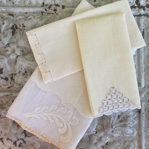 May include: A collection of ivory-colored linen napkins. One napkin features a decorative cut-out design, another has a scalloped edge with a geometric pattern, and a third has an embroidered floral design.