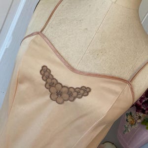 May include: A peach-colored satin slip dress with delicate spaghetti straps and a subtle floral embroidery detail. The dress features a contrasting trim along the neckline and straps, creating a vintage-inspired aesthetic.