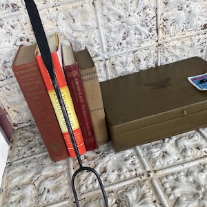 May include: A black metal fire poker with a looped handle and a two-pronged end. The poker is positioned in front of a collection of vintage books with red and brown covers. A wooden box with a decorative emblem is also visible.