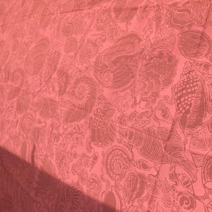May include: Coral-colored fabric with a repeating pattern of seashells in varying sizes and shapes. The design features detailed illustrations of spiral and conch shells, creating a textured appearance. The fabric is likely used for home decor or apparel.