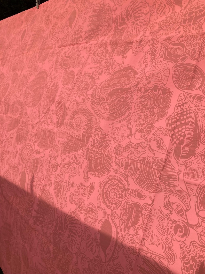 May include: Coral-colored fabric with a repeating pattern of seashells in varying sizes and shapes. The design features detailed illustrations of spiral and conch shells, creating a textured appearance. The fabric is likely used for home decor or apparel.