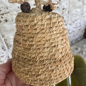 Puede incluir: Un recipiente tejido en forma de colmena de paja con una superficie texturizada. La parte superior presenta un asa de cuerda trenzada y dos elementos decorativos que se asemejan a abejas. El recipiente es de color beige natural, lo que sugiere una estética rústica o de granja.