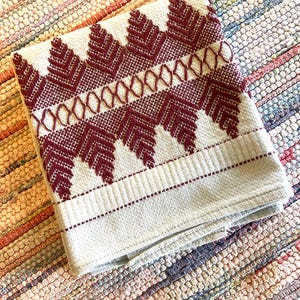May include: Folded white and burgundy hand towel with a geometric pattern. The towel features a repeating design of burgundy chevrons and a horizontal band of burgundy stitching. The towel is folded and resting on a multi-colored rug.