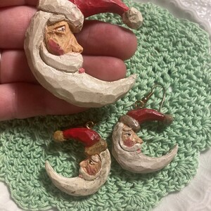 May include: A pair of Christmas earrings featuring a crescent moon with a Santa face and a red hat. The earrings are made of white and red painted wood.
