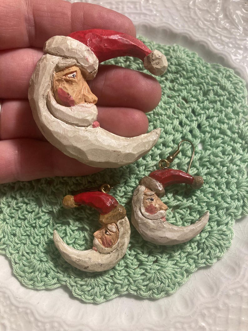 May include: A pair of Christmas earrings featuring a crescent moon with a Santa face and a red hat. The earrings are made of white and red painted wood.