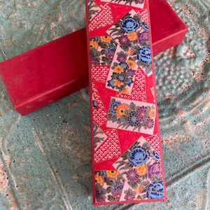 May include: A rectangular box with a lid featuring a vibrant red background decorated with a repeating pattern of pink, blue, purple, and yellow floral motifs arranged in geometric shapes, accompanied by a plain red base box underneath.