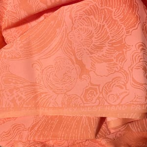 May include: Coral-colored fabric with a repeating pattern of stylized seashells and swirling lines. The fabric is folded, revealing the intricate design. The text "KEY WEST HAND PRINT FABRICS Inc" is visible.