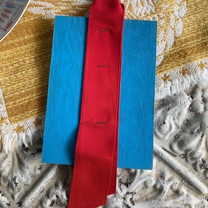 May include: A bright red ribbon with a gold ring attached, resting on a blue rectangular block. The ribbon is secured with three small stitches. The background features a textured, off-white surface and a yellow patterned cloth.