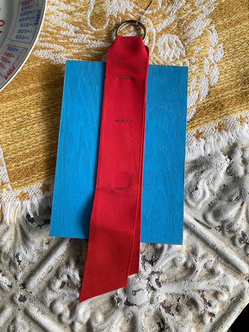 May include: A bright red ribbon with a gold ring attached, resting on a blue rectangular block. The ribbon is secured with three small stitches. The background features a textured, off-white surface and a yellow patterned cloth.