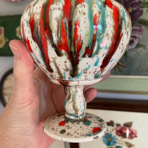 May include: A ceramic pedestal bowl with a white base and a colorful, dripped glaze design. The glaze features red, teal, and brown vertical streaks, with speckled accents. The bowl has a rounded body and a flared base.