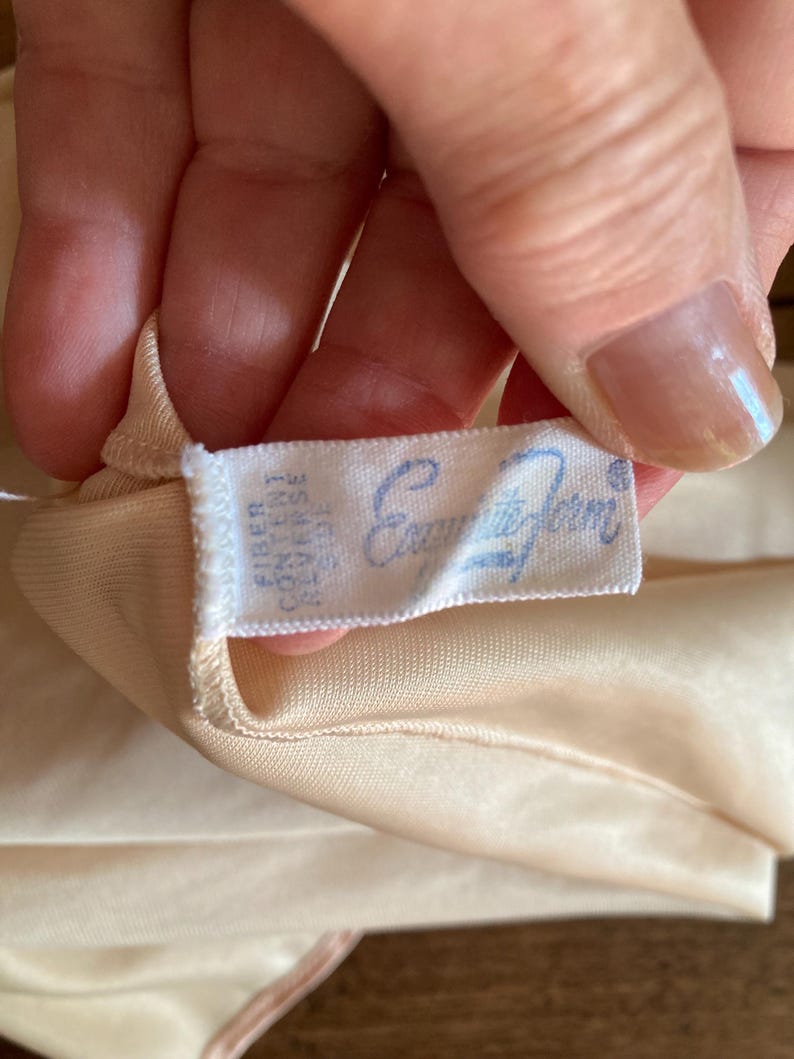 May include: Close-up of a peach-colored garment with a tag that reads "Enchant&eacute; Form" in light blue script. The fabric appears to be soft and silky, with a subtle sheen. The garment is held by a hand, showing a glimpse of the texture.