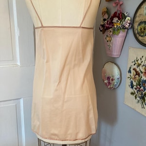 May include: A vintage peach-colored slip dress with thin spaghetti straps and a delicate trim. The slip is displayed on a mannequin, showcasing its simple, elegant design. The dress is made of a lightweight fabric, perfect for layering or wearing on its own.