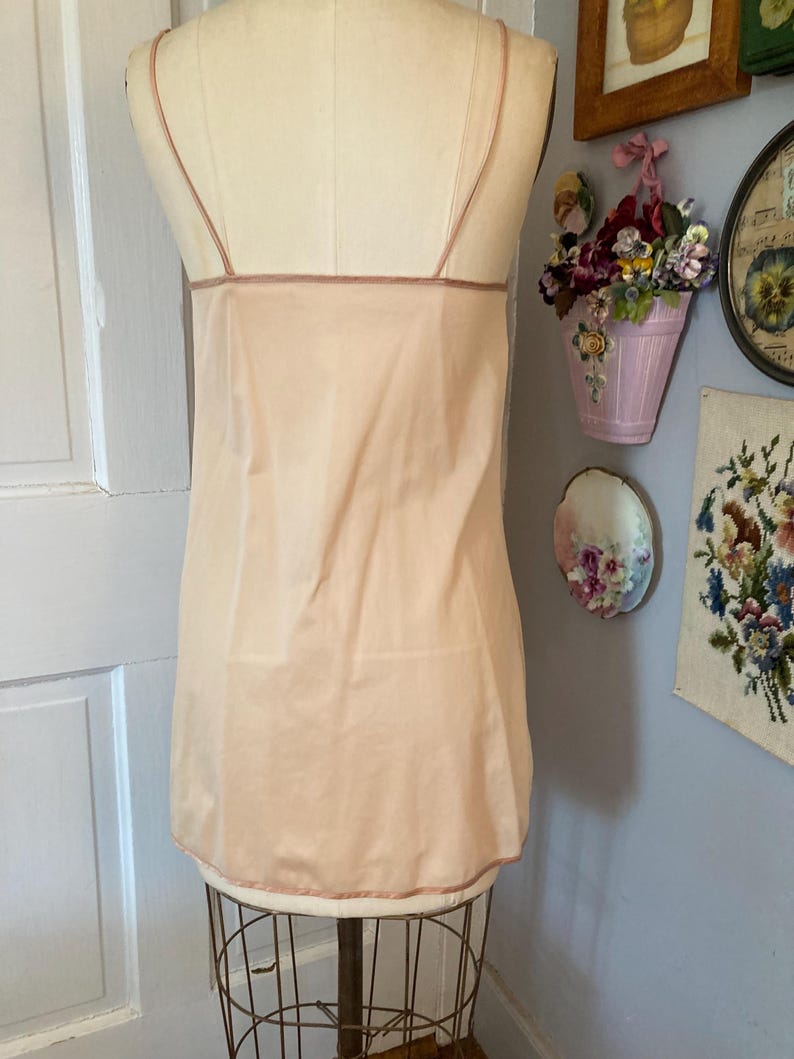 May include: A vintage peach-colored slip dress with thin spaghetti straps and a delicate trim. The slip is displayed on a mannequin, showcasing its simple, elegant design. The dress is made of a lightweight fabric, perfect for layering or wearing on its own.