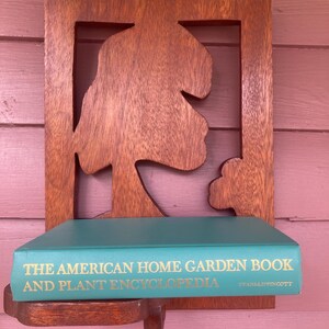 May include: A wooden wall shelf with a carved tree design. A teal book titled "The American Home Garden Book and Plant Encyclopedia" by Evans & Lippincott is on the shelf.