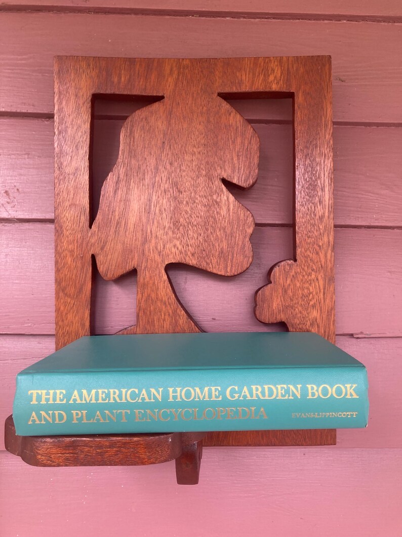 May include: A wooden wall shelf with a carved tree design. A teal book titled "The American Home Garden Book and Plant Encyclopedia" by Evans & Lippincott is on the shelf.