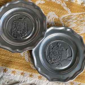 May include: Two pewter plates with a wavy edge. One plate has a drum design, and the other shows a scene with figures. The plates are on a yellow and white patterned surface.