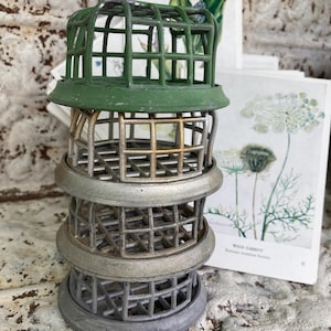 May include: A stack of four metal floral arranging cages in shades of green and silver. The cages have a grid-like design and are stacked on top of each other. Botanical art prints are in the background.