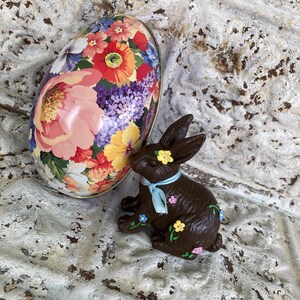 May include: A decorative Easter egg-shaped tin with a floral design and a chocolate-colored bunny figurine adorned with painted flowers and a blue ribbon. The bunny is sitting next to the egg.