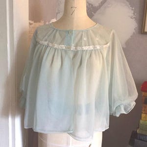 Puede incluir: Una chaqueta de cama transparente azul claro vintage con cuello redondo y mangas tres cuartos. La chaqueta presenta un delicado ribete de encaje blanco a lo largo del escote y los puños, y pequeños cierres de botón. La tela es ligera y ligeramente translúcida.