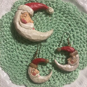 May include: A set of three wooden ornaments featuring a Santa Claus face carved into a crescent moon shape. The ornaments are painted white with red hats and have a rustic, handmade look.