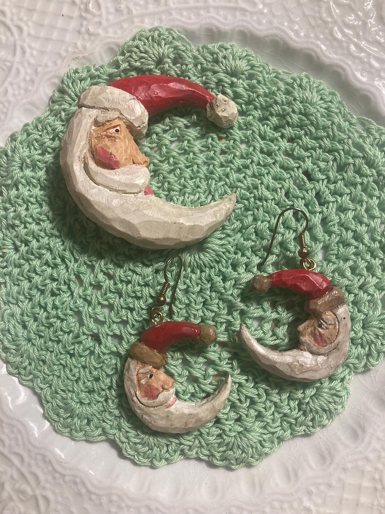 May include: A set of three wooden ornaments featuring a Santa Claus face carved into a crescent moon shape. The ornaments are painted white with red hats and have a rustic, handmade look.