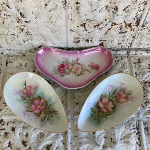 May include: Three antique porcelain dishes. A heart-shaped dish with a pink and white rim and a floral design. Two teardrop-shaped dishes with gold trim and floral designs. The dishes are arranged on a textured white surface.