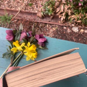 May include: A closed hardcover book with a green spine and cream colored pages. The book is lying on a blue wooden surface with a bouquet of yellow and pink flowers in the background.