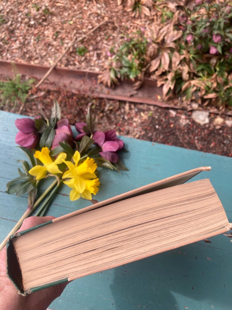 May include: A closed hardcover book with a green spine and cream colored pages. The book is lying on a blue wooden surface with a bouquet of yellow and pink flowers in the background.