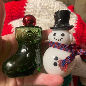 Pode incluir: Duas figuras de Natal em vidro. Uma é uma bota verde escura com uma bola vermelha. A outra é um boneco de neve branco com uma cartola preta e um cachecol vermelho, branco e azul. O boneco de neve tem olhos de pontos pretos e boca de carvão.