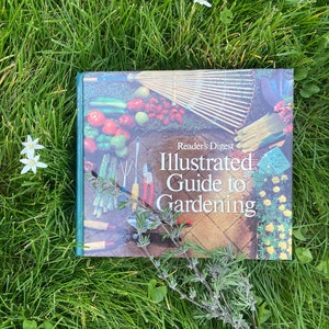 May include: A green book titled "Reader's Digest Illustrated Guide to Gardening" with a colorful image of gardening tools and plants on the cover. The book is lying on a bed of green grass with white flowers.