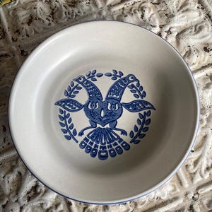 May include: A round, off-white ceramic plate with a blue rim and a central blue illustration. The illustration features a stylized bird or goat-like creature with a wreath of leaves. The plate has a slightly raised edge.