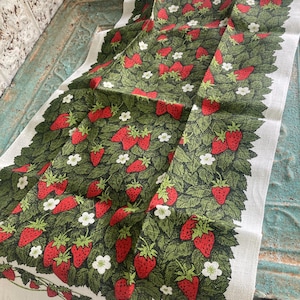 May include: A rectangular kitchen towel featuring a repeating pattern of red strawberries, green leaves, and small white flowers on a cream-colored background. The design covers the center of the fabric, framed by a decorative strawberry vine border.