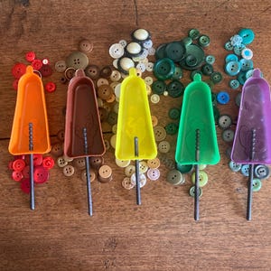 Könnte beinhalten: Fünf bunte Eis am Stiel-Formen in Orange, Braun, Gelb, Grün und Lila, jede mit einem Metallstab. Die Formen sind von einer Vielzahl von Knöpfen in Rot-, Braun-, Grün- und Blautönen auf einer Holzoberfläche umgeben.