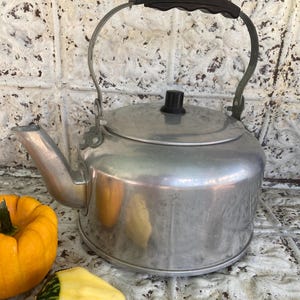 May include: A vintage aluminum kettle with a black handle and lid knob. The kettle has a spout and a curved handle. A small orange pumpkin and a yellow gourd are in the foreground. The kettle is on a tiled surface.