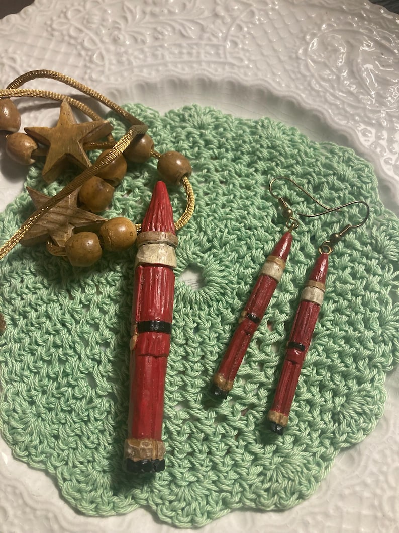 May include: A set of Christmas-themed earrings and a pendant featuring a red and white Santa Claus figure. The earrings and pendant are made of wood and painted with red, white, and black paint.