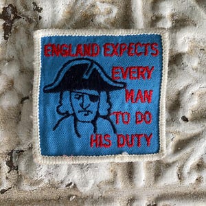 May include: A square patch with a blue background and white border. The patch features a black illustration of a man wearing a tricorn hat and an eye patch. Red text reads "ENGLAND EXPECTS EVERY MAN TO DO HIS DUTY."