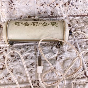 Może przedstawiać: Wintage kremowa lampa elektryczna z kwiatowym wzorem i złotym wykończeniem. Lampa ma cylindryczny kształt i stoi na dwóch małych nóżkach. Dołączony jest biały przewód i wtyczka. W tle widoczna jest miarka.