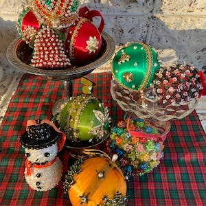 May include: A collection of colorful Christmas ornaments, including red, green, and orange spheres, a snowman, and a bell-shaped ornament. Some ornaments are adorned with beads, pearls, and gold accents. The ornaments are displayed on a plaid tablecloth and in decorative dishes.