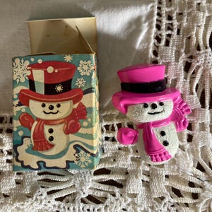 May include: A vintage snowman figurine with a pink top hat, scarf, and gloves. The snowman has a white body with black button eyes and a smiling mouth. A matching box with a snowman illustration is also present.
