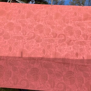 May include: Coral-colored fabric with a detailed pattern of stylized seashells and other marine life. The fabric is hanging outdoors, secured by clothespins, with a glimpse of a blue sky and trees in the background. The fabric's design is visible in the sunlight.