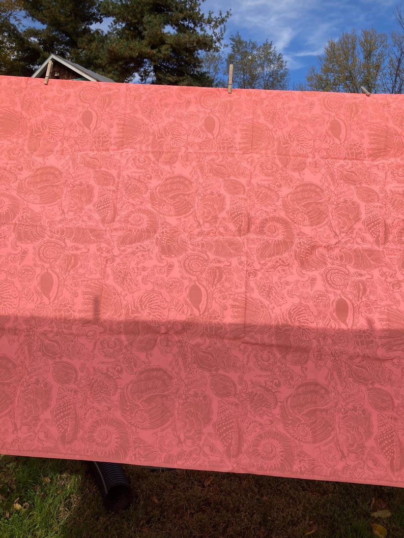 May include: Coral-colored fabric with a detailed pattern of stylized seashells and other marine life. The fabric is hanging outdoors, secured by clothespins, with a glimpse of a blue sky and trees in the background. The fabric's design is visible in the sunlight.