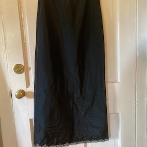 Op de afbeelding: Een zwarte onderrok hangt aan een houten kleerhanger tegen een witte deur. De rok is gemaakt van een gladde, donkere stof en heeft een delicate kanten rand aan de zoom. De rok is lang en reikt tot de enkels.