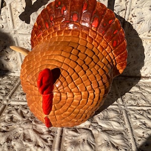 May include: A ceramic turkey ornament with a detailed, textured design. The turkey has a brown body, a red wattle, and a fan-shaped tail with reddish-brown accents. Likely a decorative item for the autumn or Thanksgiving holiday.