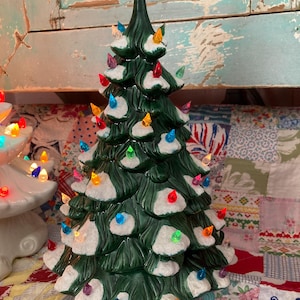 Könnte beinhalten: Ein grüner Keramik-Weihnachtsbaum, verziert mit bunten Lichtern und einem grünen Stern. Der Baum hat weiße Akzente, die Schnee simulieren. Der Sockel ist cremefarben.