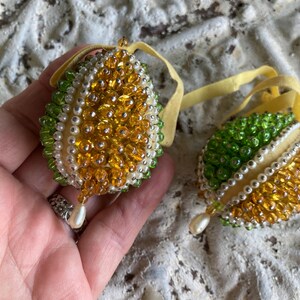 Puede incluir: Dos adornos en forma de huevo decorados con cuentas de color ámbar, verde y blanco. Cada adorno cuenta con una cinta amarilla en la parte superior y una cuenta blanca en forma de lágrima colgando de la parte inferior, creando una superficie texturizada.