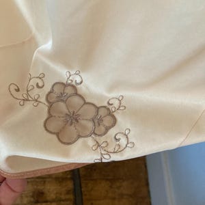 May include: Close-up of a cream-colored fabric with a floral embroidery design in shades of brown. The embroidery features a cluster of flowers and delicate vines, adding a touch of elegance to the textile.