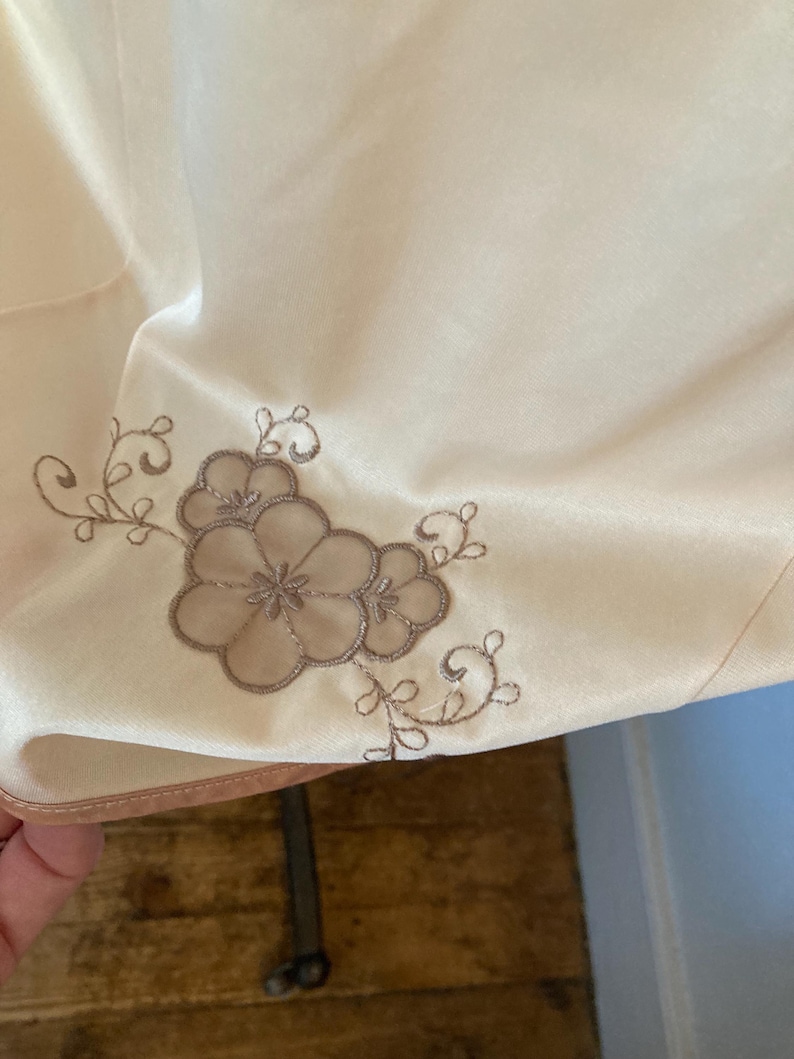 May include: Close-up of a cream-colored fabric with a floral embroidery design in shades of brown. The embroidery features a cluster of flowers and delicate vines, adding a touch of elegance to the textile.